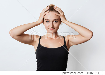 Gorgeous female athlet prepares for morning running, ties hair in bun, keeps hands on head, looks delightfully into camera. Beautiful woman has some troubles, shows it with gestures, poses in studio Gorgeous female athlet prepares for morning running, ties hair in bun, keeps hands on head, looks delightfully into camera. Beautiful woman has some troubles, shows it with gestures, poses in studio 100232413