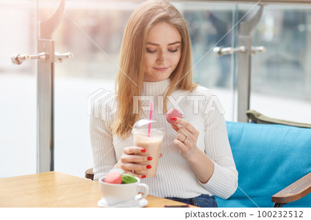 Slender sweet positive female holding plastic cup with coctail and one pink macaroon, going to have bite, looking at it attentively, sitting at table in cafeteria, wearing smart white sweater. Slender sweet positive female holding plastic cup with coctail and one pink macaroon, going to have bite, looking at it attentively, sitting at table in cafeteria, wearing smart white sweater. 100232512