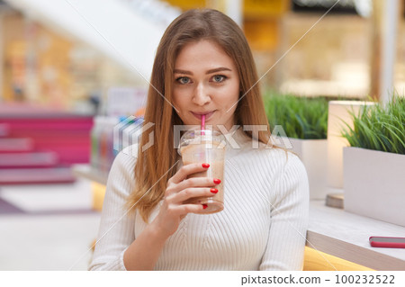 Lovely young girl looks at camera with satisfied expression, drinks milk cocktail, wears white jumper, has make up, poses against cafe blurred interior, enjoys free time. Peope, drinking concept Lovely young girl looks at camera with satisfied expression, drinks milk cocktail, wears white jumper, has make up, poses against cafe blurred interior, enjoys free time. Peope, drinking concept 100232522