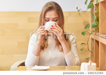 Portrait of positive calm female sipping hot drink in cafe with wooden stylish interior, holds cup in hands tightly, spends weekends in warm convenient place. Free time activities concept. Portrait of positive calm female sipping hot drink in cafe with wooden stylish interior, holds cup in hands tightly, spends weekends in warm convenient place. Free time activities concept. 100232565