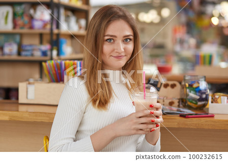 Woman drinking cocktail at bar. Pretty girl with blond hair, in white casual shirt with red fashionable manicure. Young adorable female holds plastic cup, looks at camera, enjoing her resting. Woman drinking cocktail at bar. Pretty girl with blond hair, in white casual shirt with red fashionable manicure. Young adorable female holds plastic cup, looks at camera, enjoing her resting. 100232615