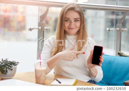 Adorable cheerful young Caucasian woman holds smart phone and points fore finger at black display, showing copyspace isolated over cafe background. Electronic gadgets, devices and people concept. 100232630