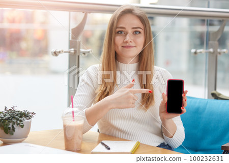 Sweet blond haired female sitting in cafe, holding smartphone, focusing on it with help of forefinger, being on lunch, having notes and plastic cup of coctail on table, looks glad. Ad concept. 100232631