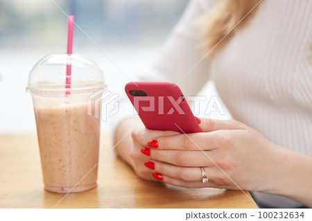 Faceless woman holds modern cell phone in hands, has red manicure, checks notification, drinks milkshake from plastic cup with straw, connected to wireless internet in cafeteria. Technology concept Faceless woman holds modern cell phone in hands, has red manicure, checks notification, drinks milkshake from plastic cup with straw, connected to wireless internet in cafeteria. Technology concept 100232634