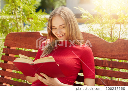 People, rest, hobby, vacation concept. Cheerful fair-haired woman in red sweater holding her favourite book reading funny stories. Pretty female reading book at wooden bench admiring weather People, rest, hobby, vacation concept. Cheerful fair-haired woman in red sweater holding her favourite book reading funny stories. Pretty female reading book at wooden bench admiring weather 100232668