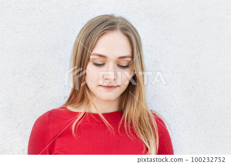 Adorable blonde woman in red dress looking thoughtfully down thinking over her relationships with boyfriend having sad expression. Young lady thinking about something while looking pensively down 100232752