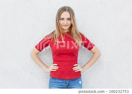 Waist up portrait of confident fair-haired woman in red sweater holding her hands on waist posing against white concrete wall. Elegant young female with attractive appearance having self-confidence 100232753