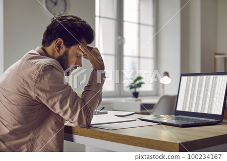 Tired, stressed man sitting at his working desk with a laptop and suffering from a headache Tired, stressed man sitting at his working desk with a laptop and suffering from a headache 100234167
