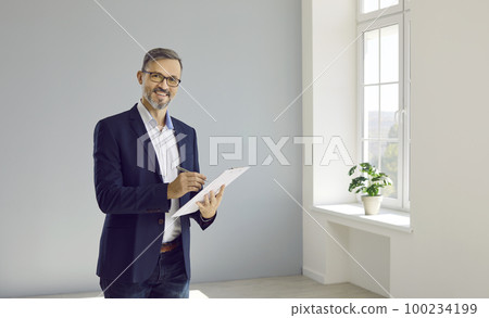 Smiling realtor man fills out documents in clipboard while standing in apartment for sale. 100234199
