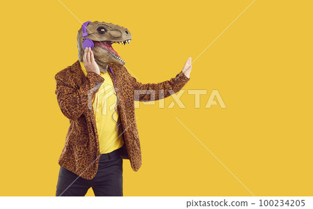 Eccentric funny showman in rubber dinosaur mask listening music in headphones on yellow background. 100234205