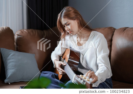 Portrait of young asian woman playing guitar on sofa relaxing stress on vacation. 100234823
