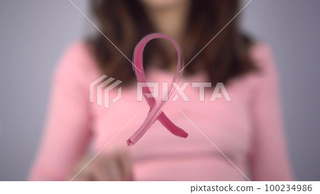 A young woman paints a breast cancer symbol on a glass. The girl draws a pink loop on the glass with a brush. 100234986