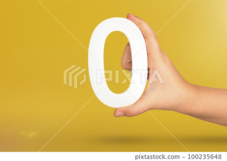Numeral zero in hand. A hand holds a white number zero on a yellow background with copy space. Zero concept, 0 percent interest rate, minimum air emissions, cost or credit no increase. Numeral zero in hand. A hand holds a white number zero on a yellow background with copy space. Zero concept, 0 percent interest rate, minimum air emissions, cost or credit no increase. 100235648
