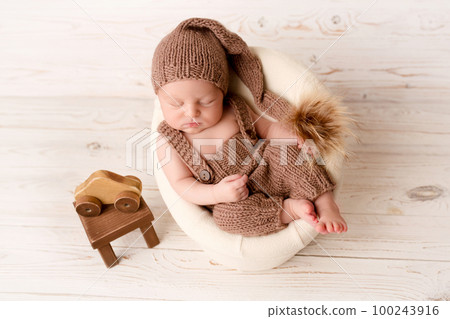 Top view of a newborn baby sleeping in a brown overalls in a brown knitted cap, in a white felt chair. On a light wooden background. Beautiful portrait of a little newborn 7 days, one week. 100243916