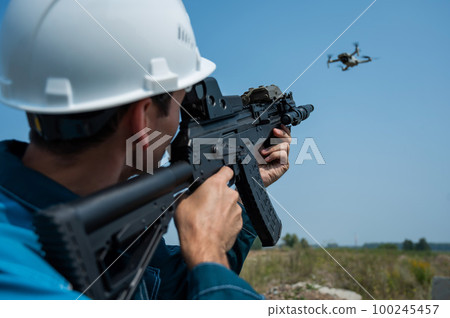 Caucasian man in a helmet shoots a flying drone with a rifle. Caucasian man in a helmet shoots a flying drone with a rifle. 100245457