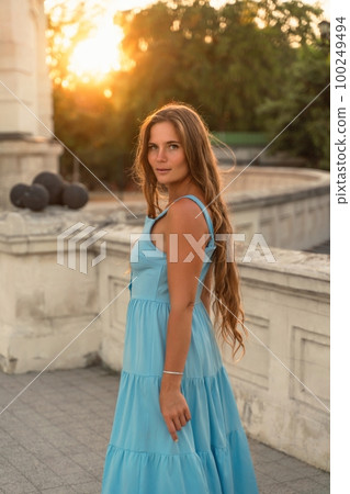 Woman sunset blue dress. Portrait of a woman with long hair and a blue dress against the backdrop of the setting sun and a white building. Lifestyle, walking around the city 100249494