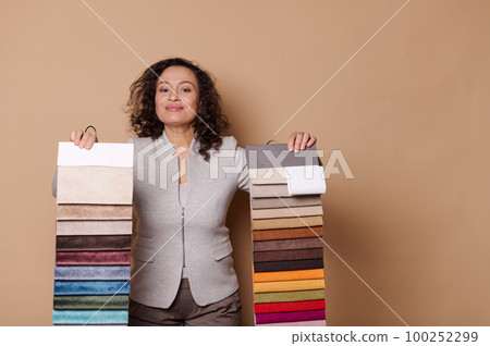 Charming middle-aged woman, interior designer, home decorator, clerk in furniture store, showing you fabric samples and various sofa upholstery materials, isolated beige background. Textile industry 100252299