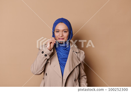 Attractive middle-aged Middle Eastern Muslim woman in blue hijab and casual beige coat, putting off her stylish spectacles, looking confidently at camera, isolated cream background. People Lifestyle Attractive middle-aged Middle Eastern Muslim woman in blue hijab and casual beige coat, putting off her stylish spectacles, looking confidently at camera, isolated cream background. People Lifestyle 100252300