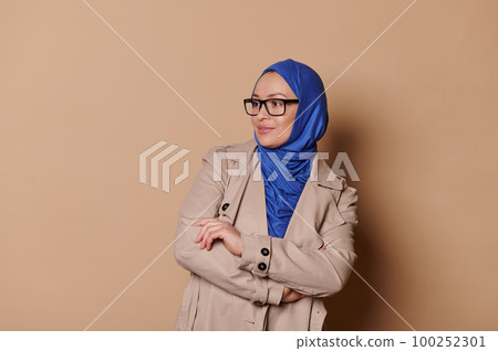 Confident successful elegant modern Muslim woman, wearing stylish eyeglasses, blue hijab and beige coat, looking to the side, poses with her arms folded over beige isolated background. Islamic fashion Confident successful elegant modern Muslim woman, wearing stylish eyeglasses, blue hijab and beige coat, looking to the side, poses with her arms folded over beige isolated background. Islamic fashion 100252301