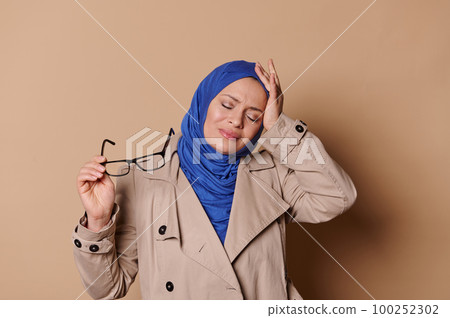 Beautiful Arab Muslim woman in blue hijab and classic stylish beige coat, putting off her spectacles, grimacing in pain, holding hand on her head, suffering from migraine, isolated on cream background 100252302