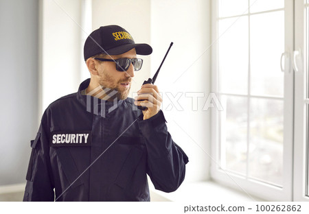 Security guard in uniform standing inside house, looking out window, and talking on radio 100262862