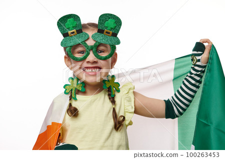 Adorable little girl wearing stylish carnival eyewear and clover leaves earrings, for Saint Patrick's Day, smiling with a cheerful toothy smile, posing with Irish flag on isolated white background Adorable little girl wearing stylish carnival eyewear and clover leaves earrings, for Saint Patrick's Day, smiling with a cheerful toothy smile, posing with Irish flag on isolated white background 100263453