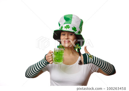 Lovely middle-aged woman in green and white striped gloves and Leprechaun's hat with clover leaves, gestures with thumb up, holding a mug with green beer, smiling looking at camera, white background Lovely middle-aged woman in green and white striped gloves and Leprechaun's hat with clover leaves, gestures with thumb up, holding a mug with green beer, smiling looking at camera, white background 100263458