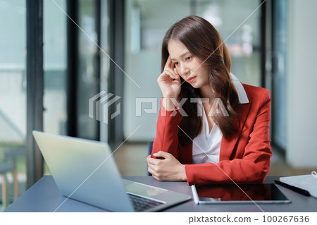 A portrait of a beautiful Asian female employee showing a stressed face while using the phone and financial documents on her desk. 100267636