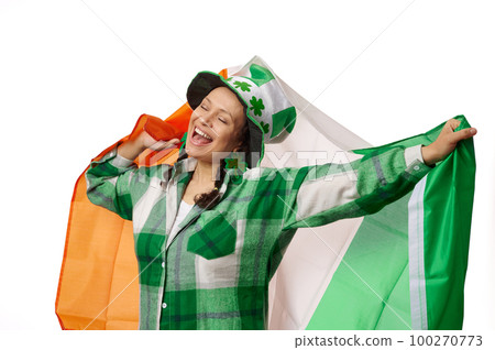 Happy woman dressed in green checkered shirt and carnival hat with clover leaves, smiling and carrying Irish flag, celebrating Saint Patrick's Day on isolated white background. Culture and traditions Happy woman dressed in green checkered shirt and carnival hat with clover leaves, smiling and carrying Irish flag, celebrating Saint Patrick's Day on isolated white background. Culture and traditions 100270773