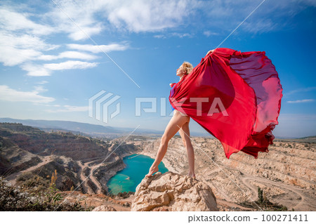 woman red dress lake mountains. Side view of a woman in a long red dress posing on a rock high above the lake. Against the background of the blue sky and the lake in the form of a heart woman red dress lake mountains. Side view of a woman in a long red dress posing on a rock high above the lake. Against the background of the blue sky and the lake in the form of a heart 100271411