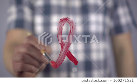 A young man paints with a brush the symbol of hiv in red. A man draws a red ribbon loop on the glass in support of cancer patients. Close-up. 100271551