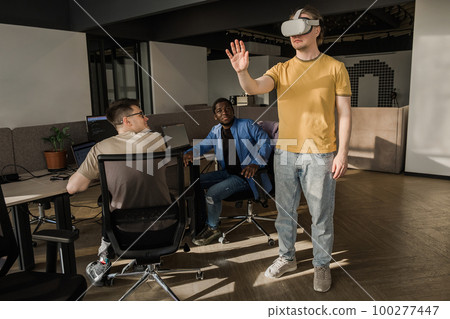 IT Development Team Working on VR Project. Man wearing VR headset, playing and touching something in the virtual world. Diverse men programmers using VR technologies and computer to create video game. 100277447