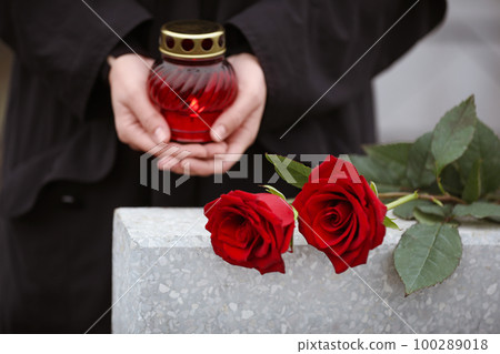 Woman holding candle near light grey tombstone with red roses outdoors, closeup. Funeral ceremony Woman holding candle near light grey tombstone with red roses outdoors, closeup. Funeral ceremony 100289018
