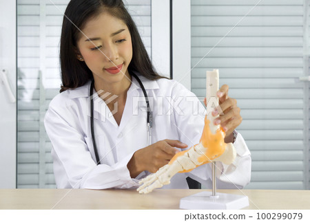 Young asian doctor in white gown and stethoscope pointing at human skeleton foot ankle bone joint anatomy model, present to patient via wireless communication. Healthcare and medicine concept. 100299079