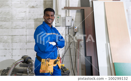 Young technician in blue mechanic jumpsuit, protective glove and yellow Tool Belts stand with arms crossed in a wooden furniture factory. 100299132
