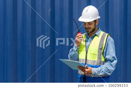 Shipment worker with safety vest and hardhat looking at clipboard while holding walkie talkie. A large steel cargo container is in the background. Shipment worker with safety vest and hardhat looking at clipboard while holding walkie talkie. A large steel cargo container is in the background. 100299153