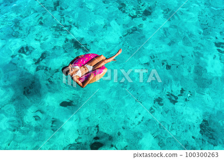 Beach vacation woman relaxing in pool float donut inflatable ring floating on turquoise ocean water background in Caribbean travel summer. Girl in white bikini top drone view 100300263