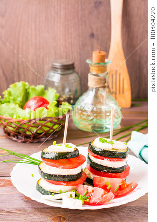 Original appetizer from fried zucchini, fresh tomatoes and feta cheese on a wooden skewer on a white plate 100304220