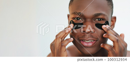 Eye patches. Facial treatment. Anti-aging skincare. Closeup portrait of guy applying black moisturizing face skin gel pads cosmetic product on light empty space background. 100312934