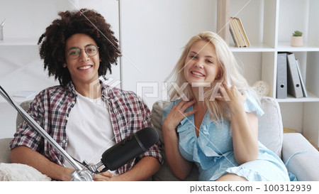 Talk show. Couple podcast. Communication stream. Cheerful smiling multiethnic influencers recording conversation using microphone at home studio interior. 100312939