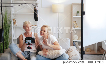 Family vlog. Influencer lifestyle. Relaxed mother daughter on couch speaking on camera on tripod in light equipped home studio interior. 100312948