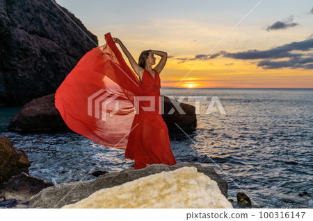 Woman in a red flying dress on the ocean or on the sea beach against the backdrop of the sunset sky. 100316147
