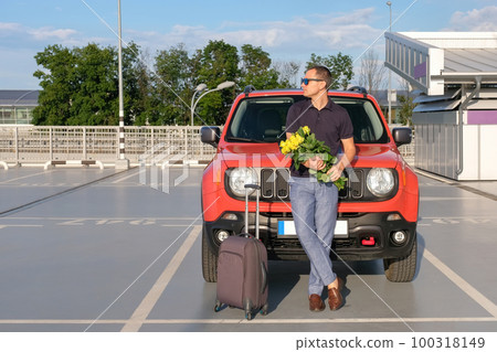 A young man with a bouquet of flowers and a suitcase stands outside his car outdoors A young man with a bouquet of flowers and a suitcase stands outside his car outdoors 100318149