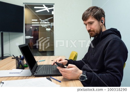 Young man working in an office sitting at his desk. Man using smartphone in the workplace Young man working in an office sitting at his desk. Man using smartphone in the workplace 100318290