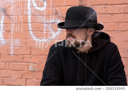 Young man in black clothes smoking a cigarette outdoors against a brick wall. 100319343