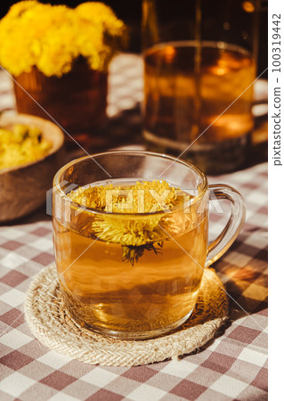 Dandelion flower healthy tea in glass teapot and glass cup on table. Delicious herbal Hot tea from fresh dandelion flowers at home at summer day. Green clearing. Bouquet of dandelions petals 100319442