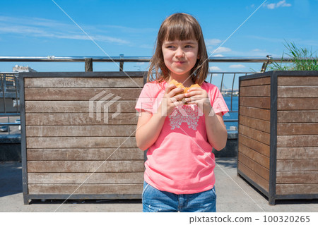 A charming smiling little girle is holding a hamburger in the open air on a sunny day A charming smiling little girle is holding a hamburger in the open air on a sunny day 100320265