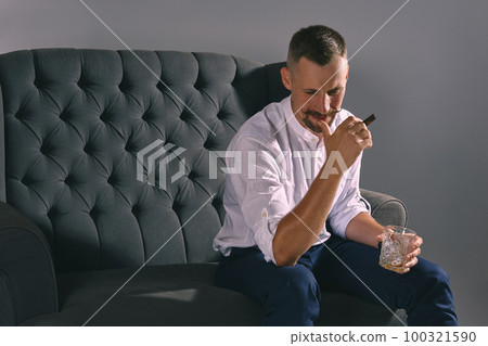 Man with stylish mustache, dressed in white shirt, blue trousers is sitting on dark sofa, holding glass of whiskey, cigar. Grey background, close-up. Man with stylish mustache, dressed in white shirt, blue trousers is sitting on dark sofa, holding glass of whiskey, cigar. Grey background, close-up. 100321590
