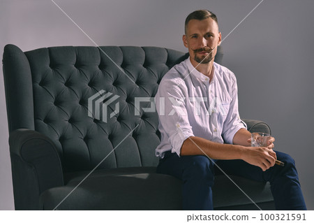 Man with stylish mustache, dressed in white shirt, blue trousers is sitting on dark sofa, holding glass of whiskey, cigar. Grey background, close-up. Man with stylish mustache, dressed in white shirt, blue trousers is sitting on dark sofa, holding glass of whiskey, cigar. Grey background, close-up. 100321591