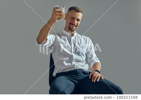Man with stylish mustache, in classic white shirt and blue trousers is sitting on chair, holding glass of whiskey. Grey background, close-up shot. Man with stylish mustache, in classic white shirt and blue trousers is sitting on chair, holding glass of whiskey. Grey background, close-up shot. 100321598